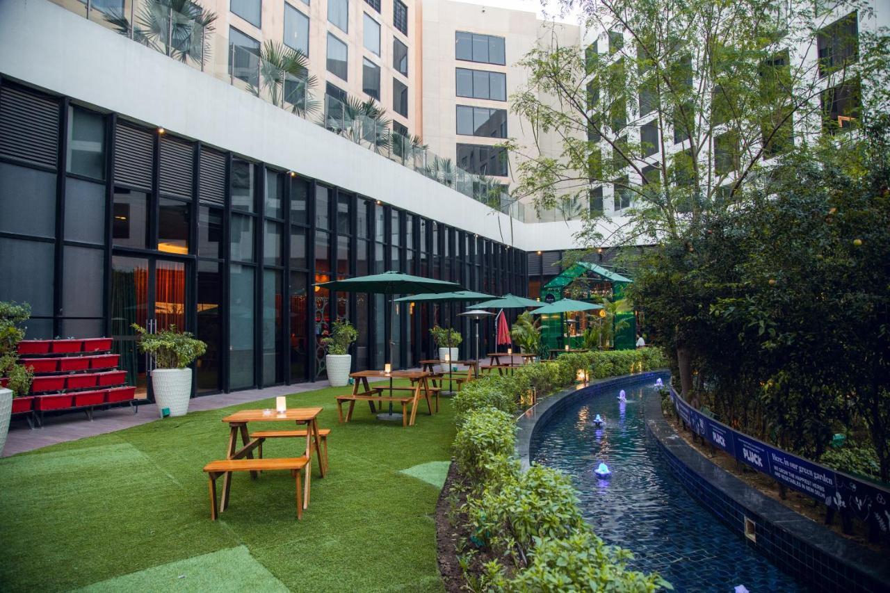 Novotel New Delhi Aerocity Buitenkant foto The photo shows a modern outdoor space that appears to be part of a hotel or a recreational area. There are several wooden tables with benches arranged on a green lawn, suggesting a place for guests to relax or dine outdoors. In the background, there