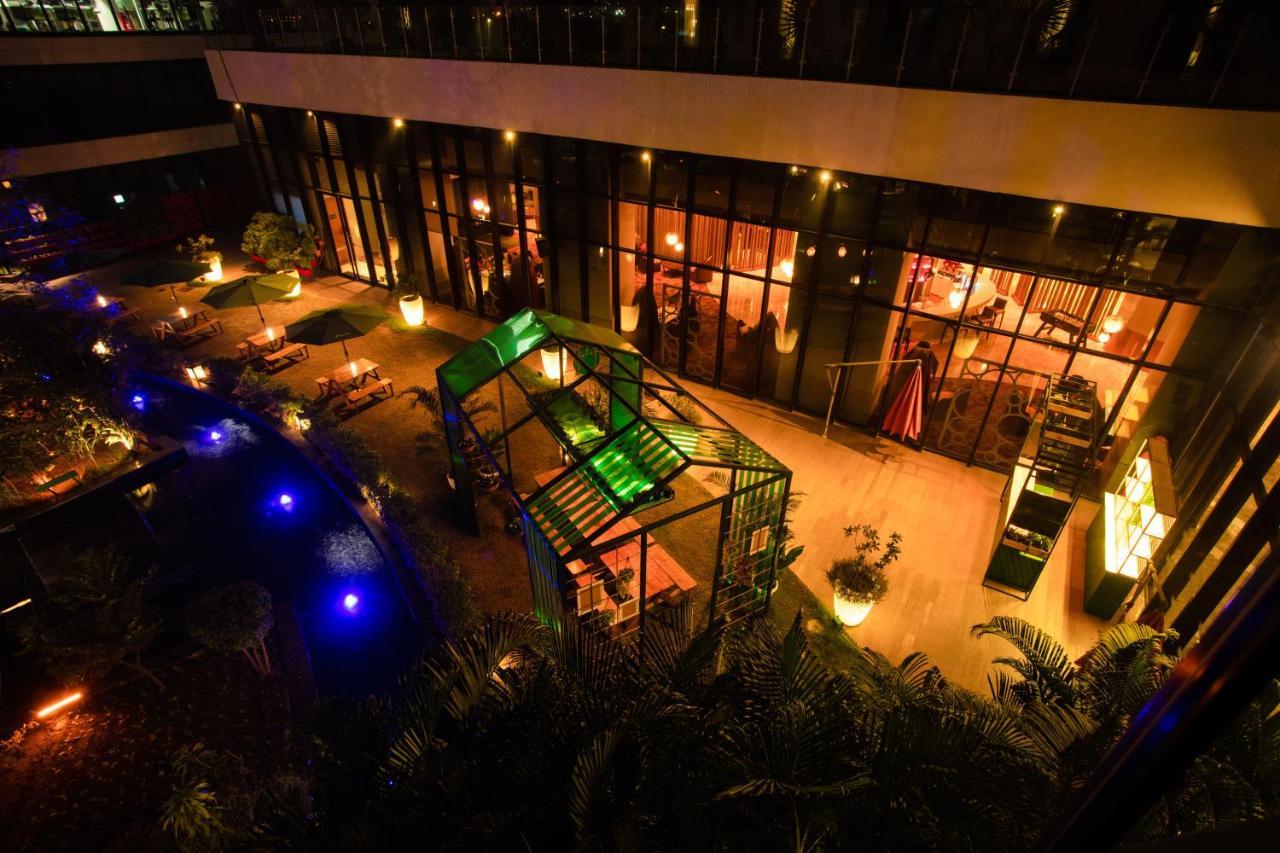 Novotel New Delhi Aerocity Buitenkant foto The photo shows an outdoor area of a restaurant or café at night. The setting features a well-lit space with various seating arrangements, surrounded by greenery. In the center, there is a striking structure, possibly a gazebo, that is illuminated wi