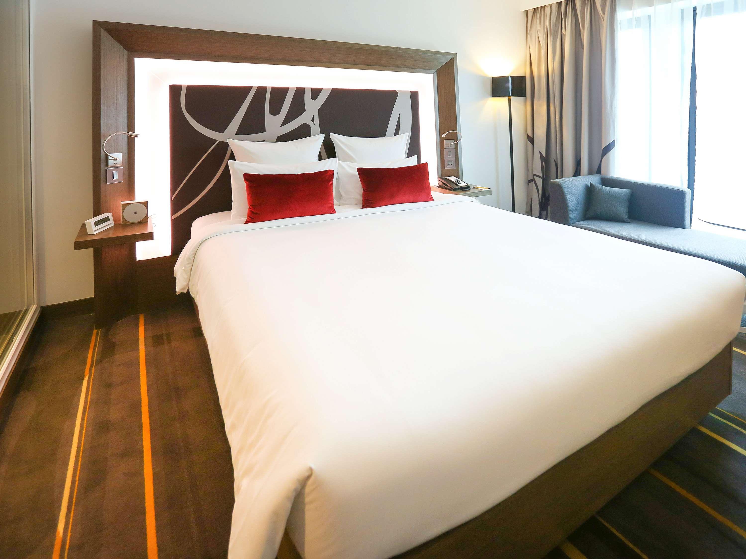 Novotel New Delhi Aerocity Buitenkant foto The photo shows a modern hotel room featuring a large bed with a white comforter. The bed has two decorative pillows in red. Behind the bed is a stylish headboard that has a contemporary design, and the room is well-lit with natural light coming from