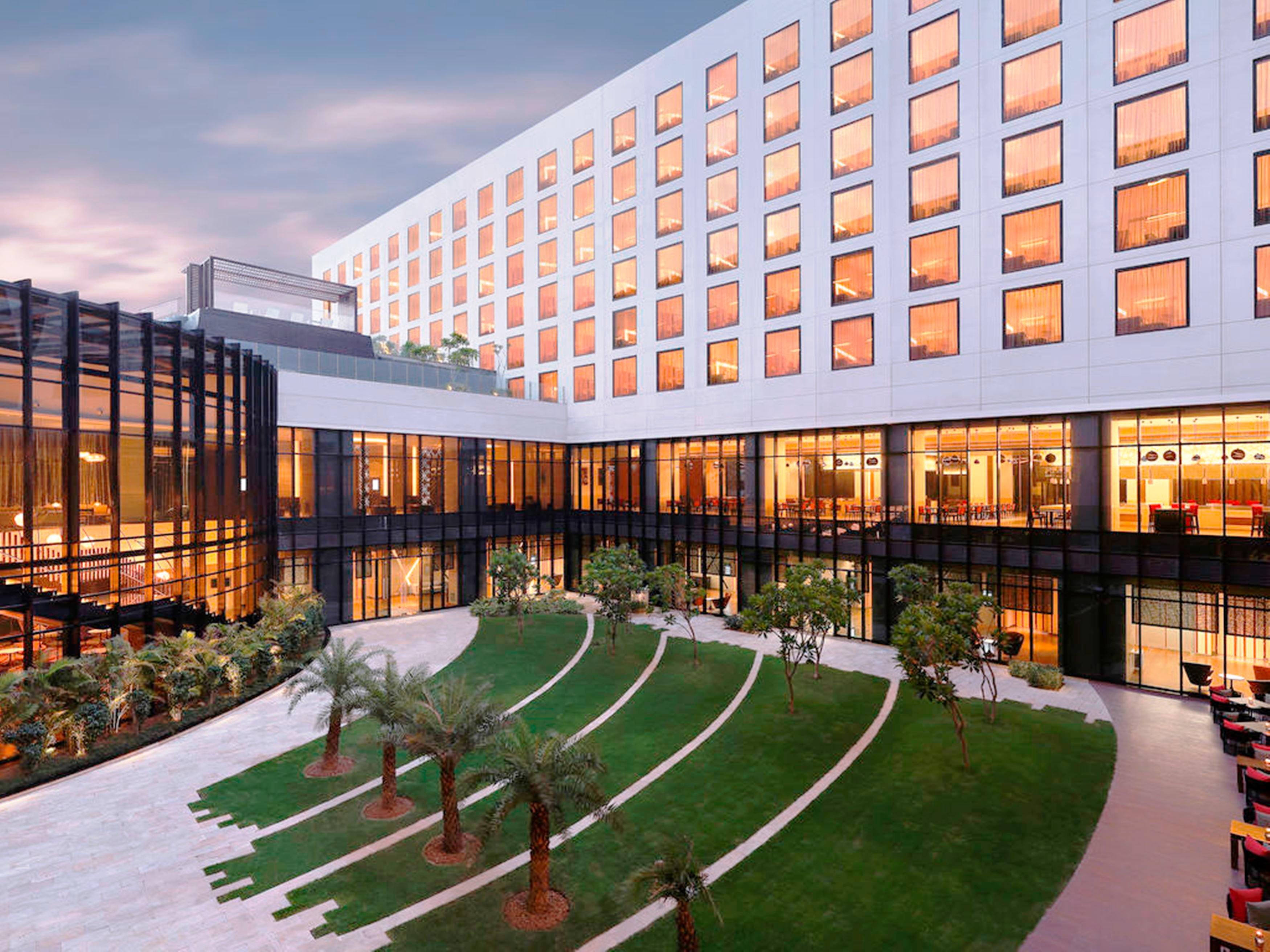 Novotel New Delhi Aerocity Buitenkant foto The photo shows a modern hotel or building complex with a sleek architectural design. In the foreground, there is a well-maintained green lawn featuring landscaped areas and small trees. The building has large glass windows that reflect the sky, and 