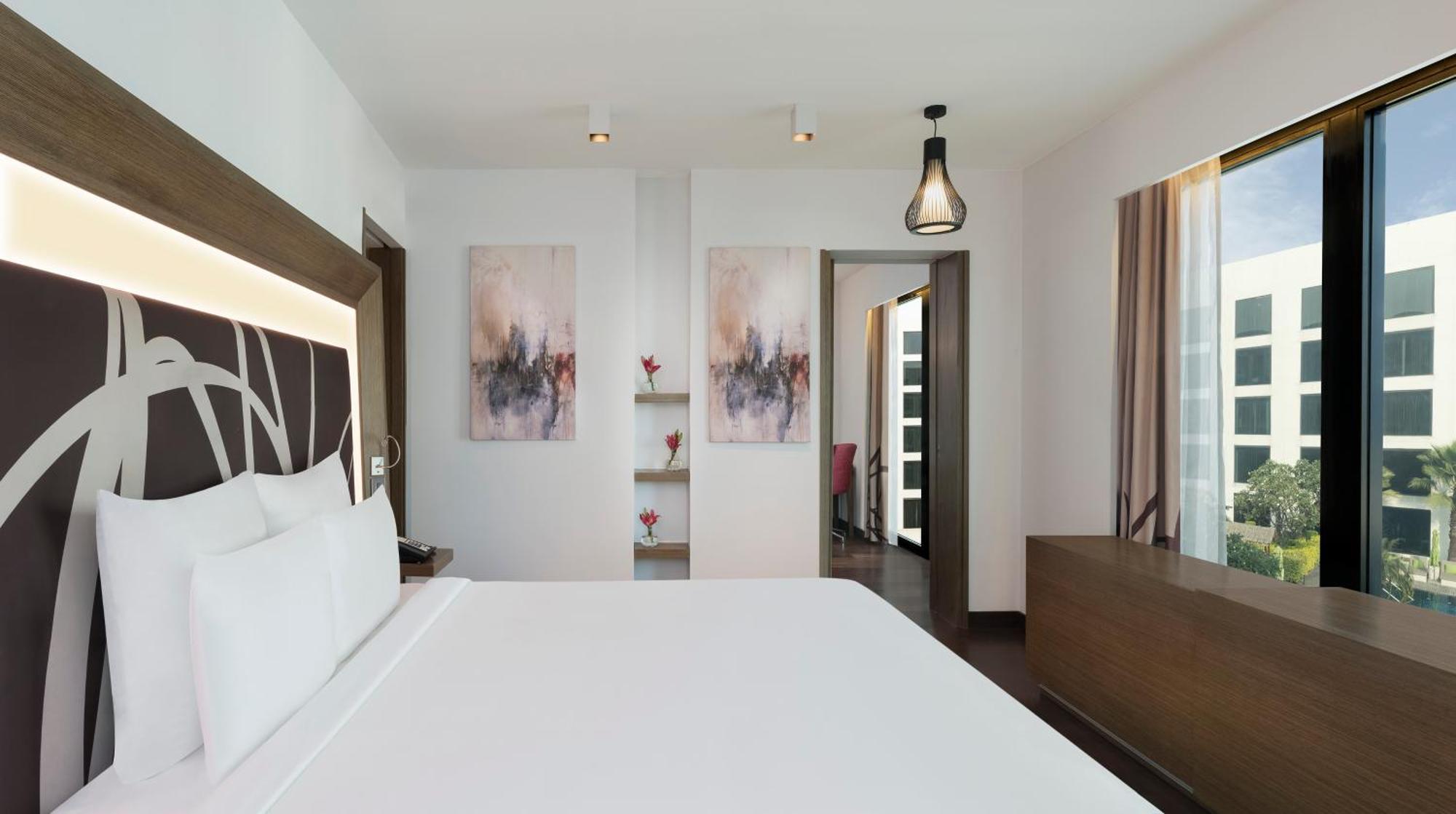 Novotel New Delhi Aerocity Buitenkant foto The photo shows a modern hotel room featuring a large white bed with a decorative headboard. The room has a clean and contemporary design, highlighted by two abstract artworks hanging on the wall above a shelf with small decorative flowers. There are