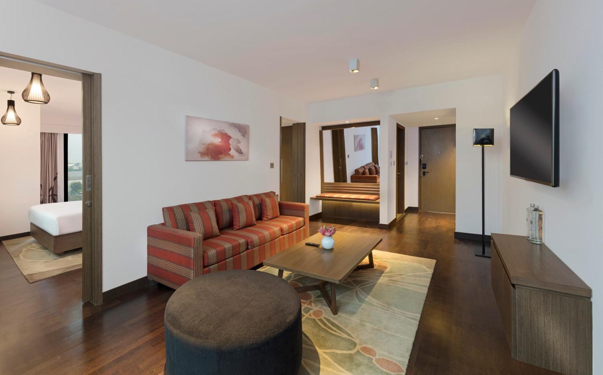 Novotel New Delhi Aerocity Buitenkant foto The photo shows a modern hotel suite or living area. It features a striped sofa with cushions, a round coffee table, and a decorative rug on a wooden floor. There is a flat-screen TV mounted on the wall, and a small side lamp nearby. The layout inclu
