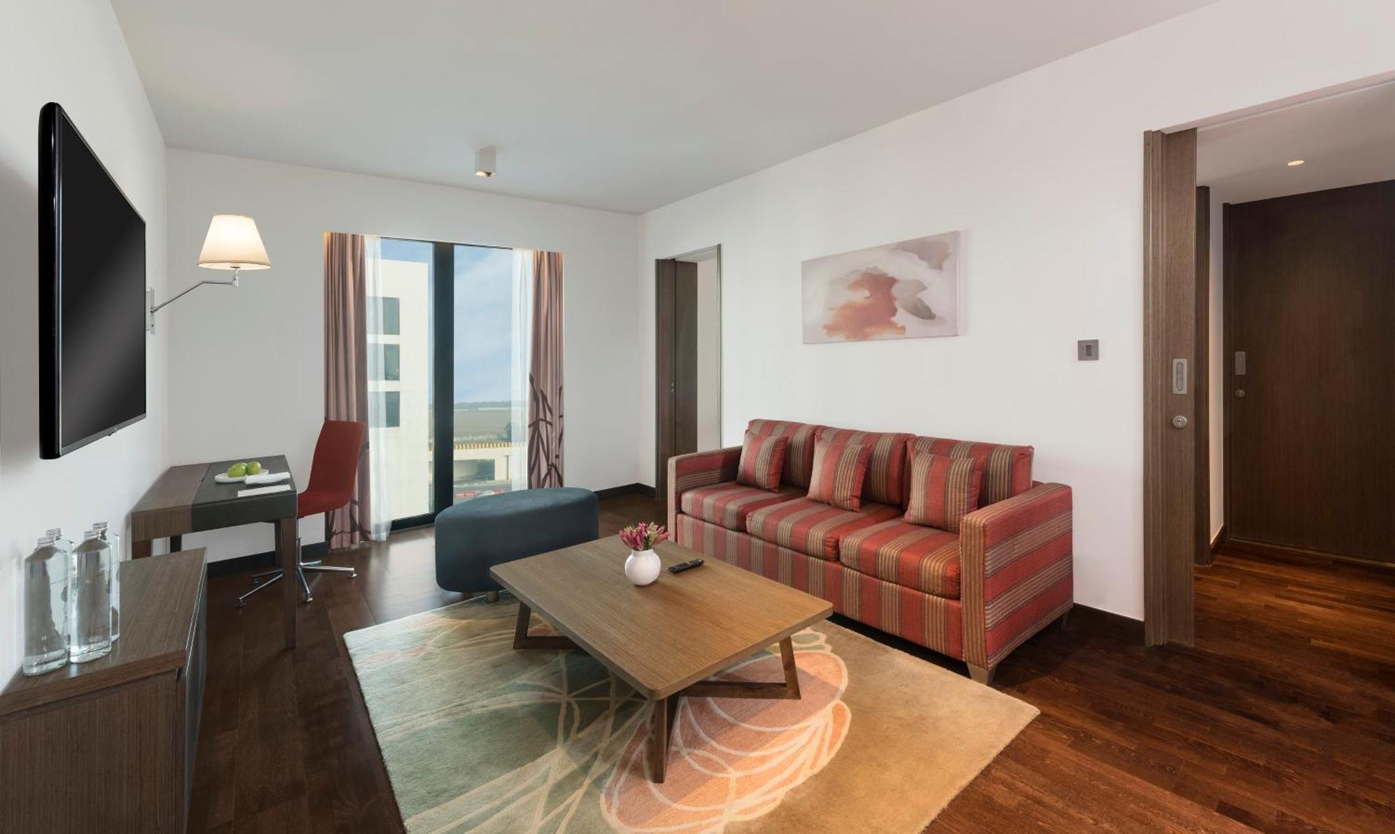 Novotel New Delhi Aerocity Buitenkant foto The photo shows a modern living room with a contemporary design. It features a striped sofa in shades of red and brown, positioned near a coffee table. The coffee table is dark in color and has a small decorative vase on it. There’s a light gray area