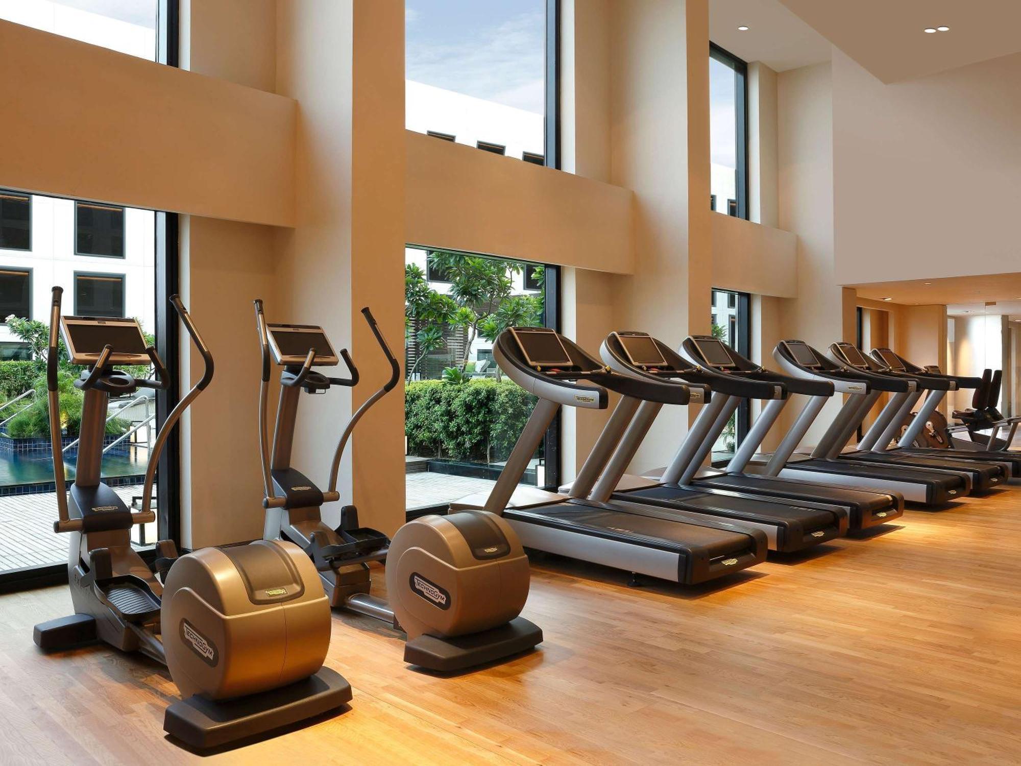 Novotel New Delhi Aerocity Buitenkant foto The photo shows a modern gym area featuring several pieces of workout equipment. On the left side, there are elliptical machines and stationary bikes, while the right side has a row of treadmills. The gym has large windows that allow natural light to