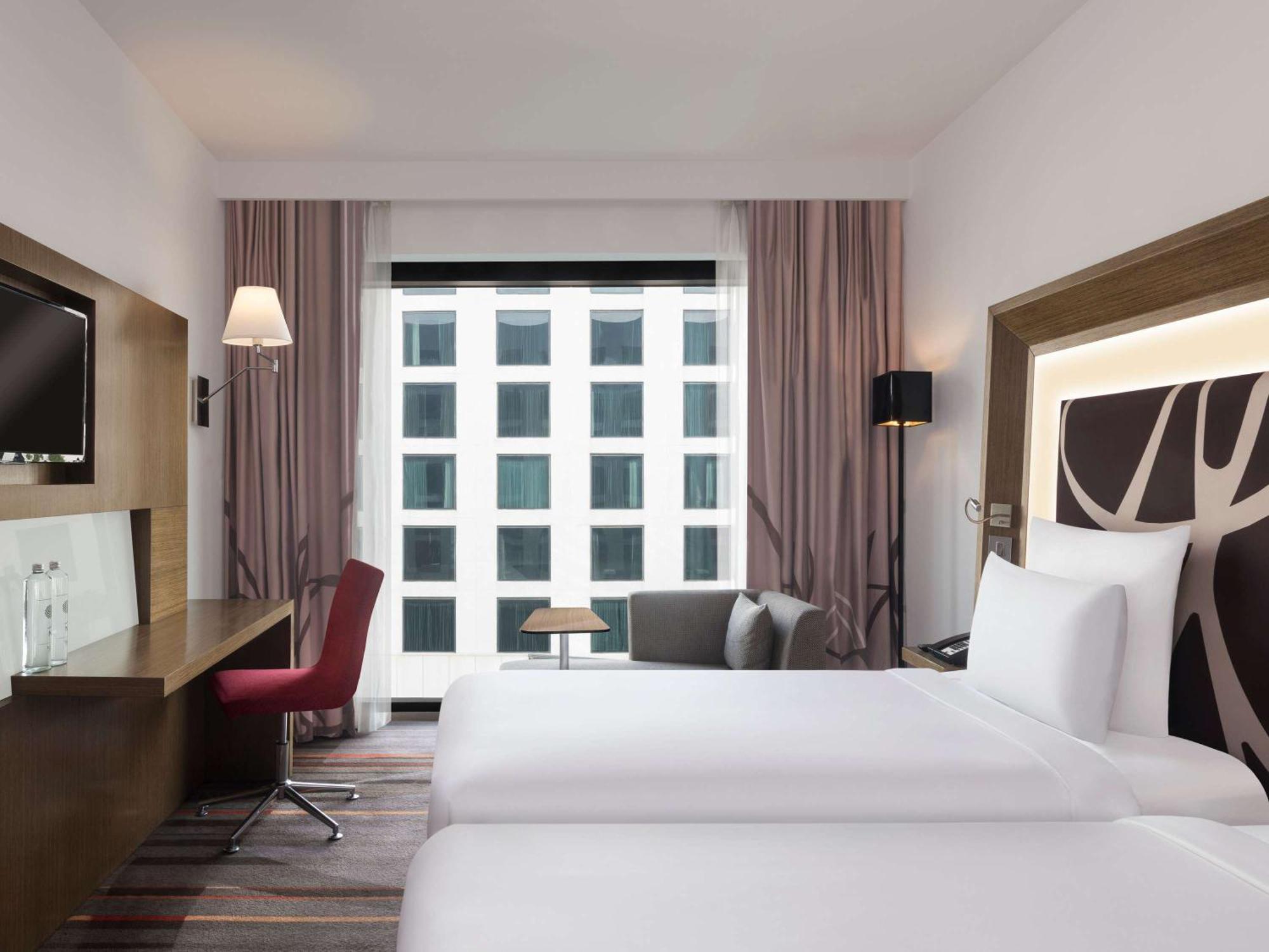 Novotel New Delhi Aerocity Buitenkant foto The photo shows a modern hotel room featuring two single beds with white linens. There is a wooden headboard on one side, and the room is designed with neutral colors. A large window is present, allowing natural light to enter and providing a view of