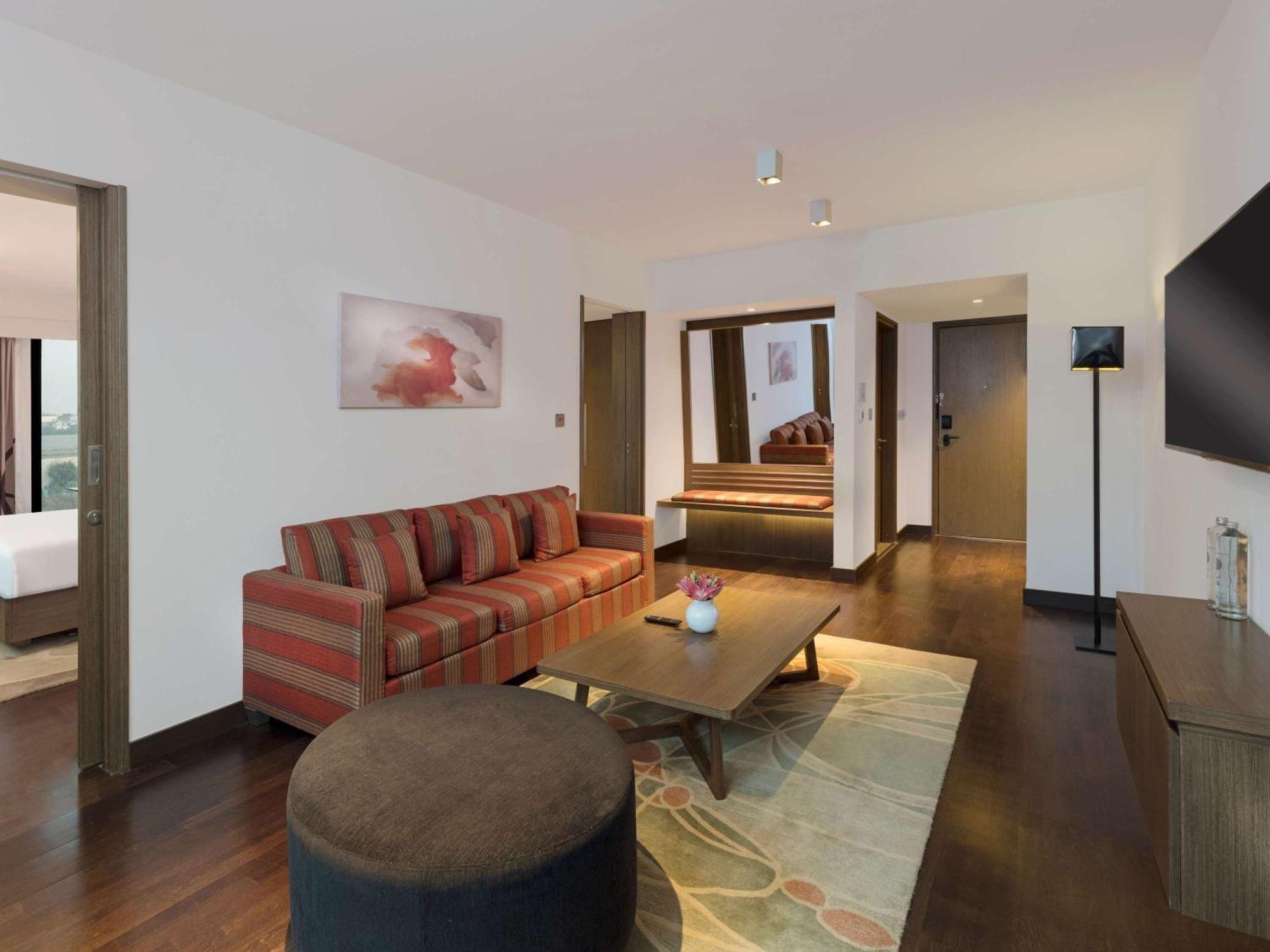 Novotel New Delhi Aerocity Buitenkant foto The photo shows a modern hotel room or suite. It features a spacious living area with a striped sofa and a round ottoman. There is a wooden coffee table in the center, and a decorative rug on the floor. The walls are painted in light colors, and ther