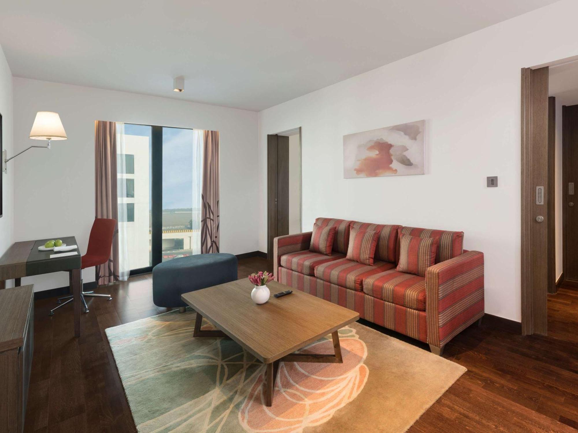 Novotel New Delhi Aerocity Buitenkant foto The photo shows a modern living room area. It features a striped sofa with red and beige tones, arranged facing a coffee table. The coffee table is wooden and has a decorative vase in the center. There's a large area rug underneath, adding texture to
