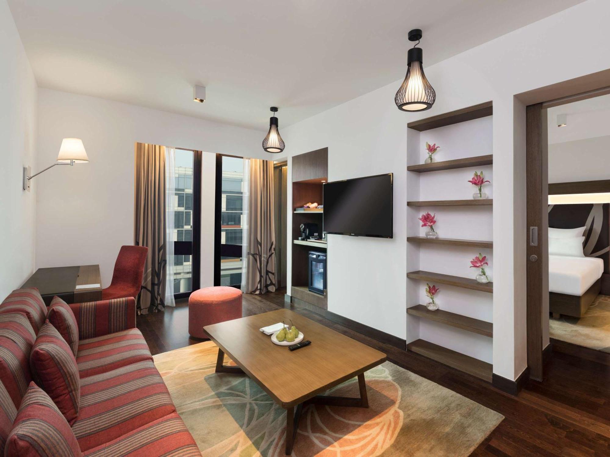 Novotel New Delhi Aerocity Buitenkant foto The photo shows a modern hotel suite or living area. It features a cozy seating arrangement with a striped sofa and an accent chair, positioned around a wooden coffee table. The decor includes two hanging pendant lights and a large flat-screen televi