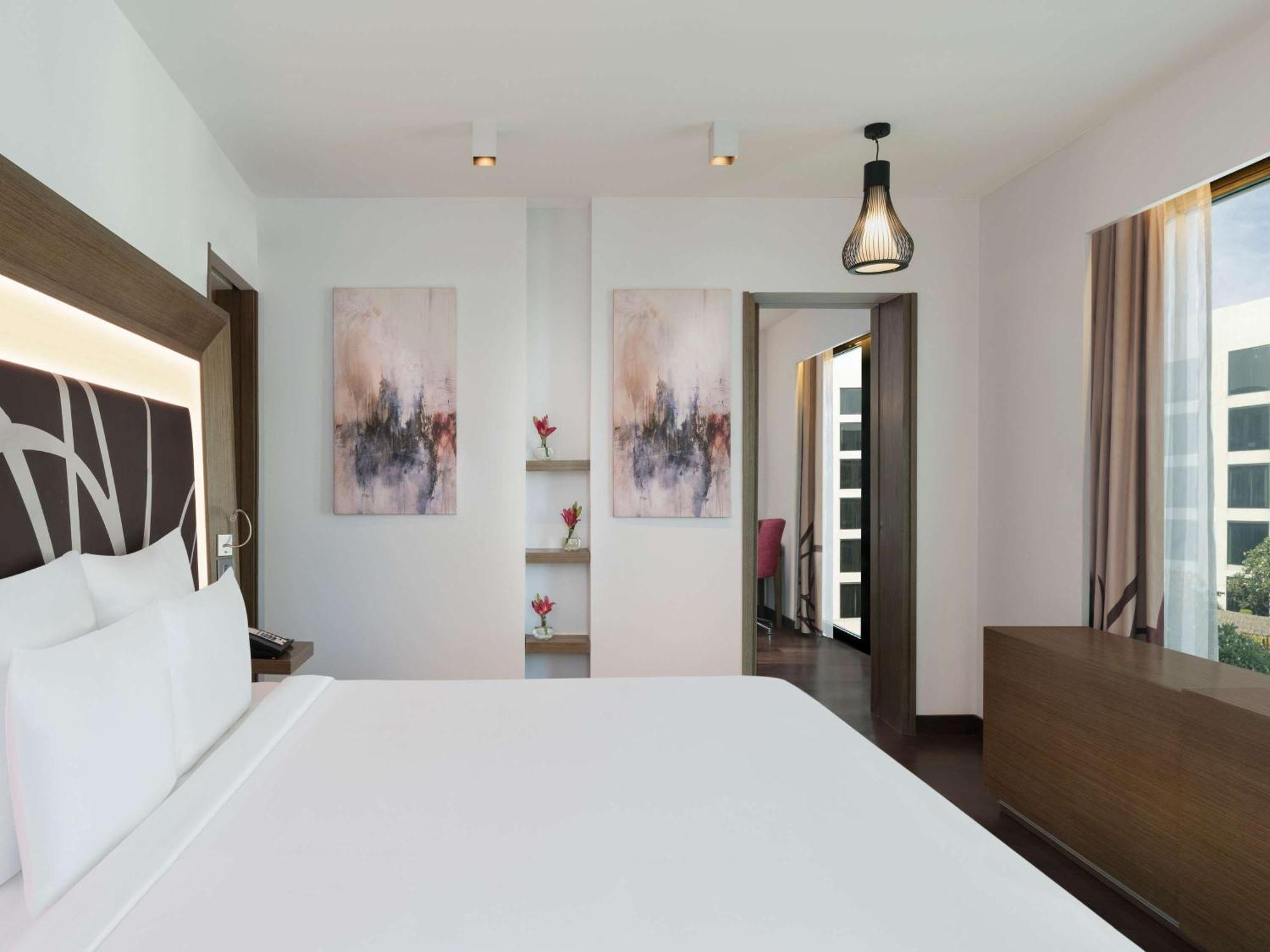 Novotel New Delhi Aerocity Buitenkant foto The photo shows a modern and stylish hotel room. The room features a large bed with a white bedding set, along with a headboard that has a contemporary design. On the walls, there are two abstract paintings that add a touch of color and elegance. To 