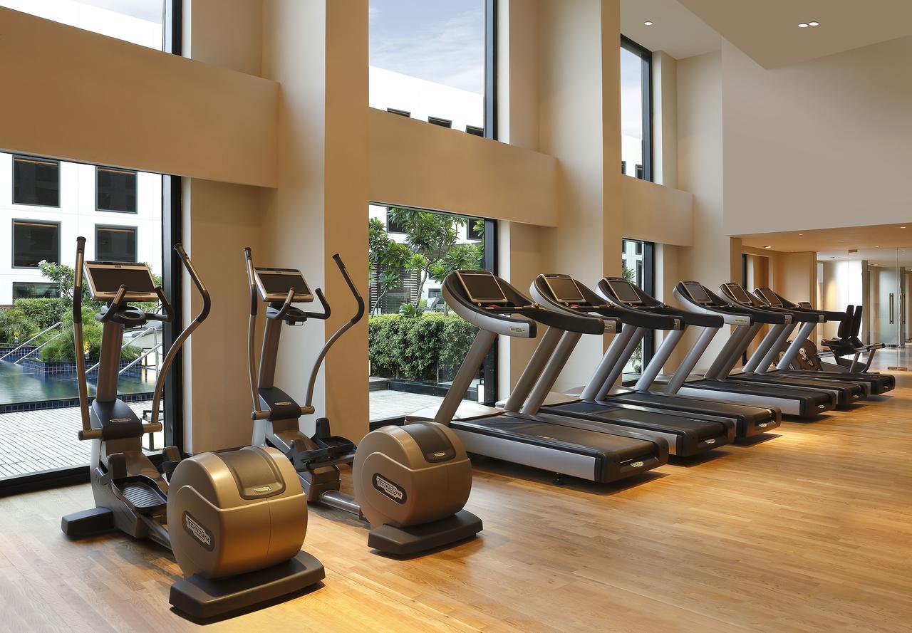 Novotel New Delhi Aerocity Buitenkant foto The photo shows a modern gym or fitness area featuring several pieces of exercise equipment. On the left, there are elliptical machines, and on the right, there are multiple treadmills lined up against large windows that allow natural light to fill t