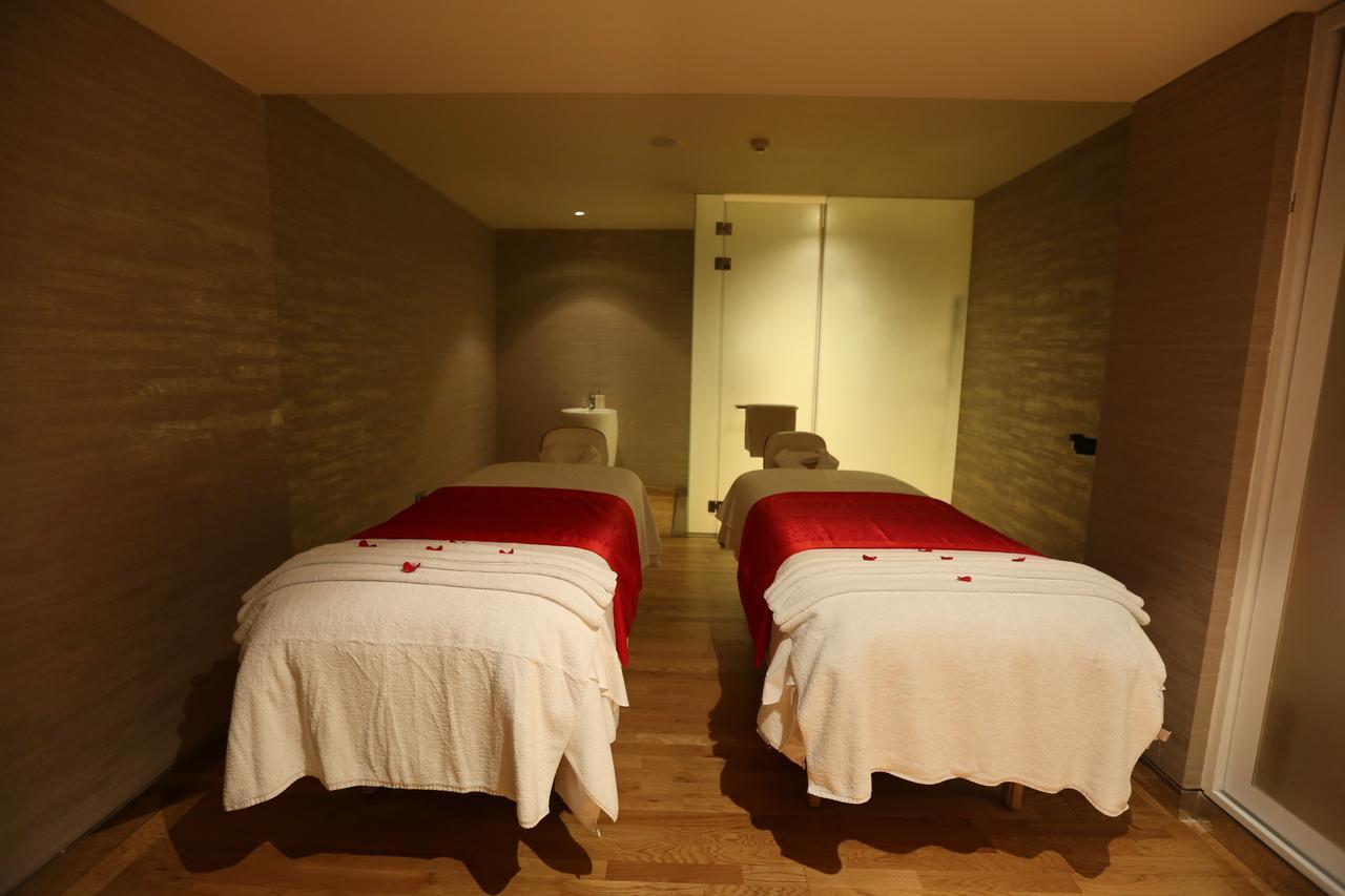 Novotel New Delhi Aerocity Buitenkant foto The photo shows a serene spa or massage room featuring two massage tables. Each table is covered with white linens, and one of them is accented with a red blanket. The room has a calming ambiance, with soft lighting and simple, neutral-toned walls. T