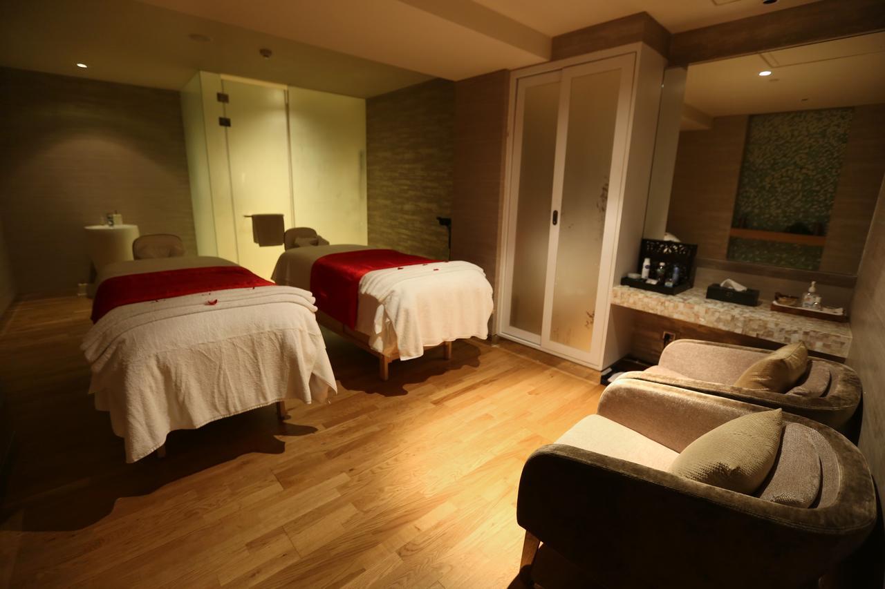 Novotel New Delhi Aerocity Buitenkant foto The photo shows a relaxing spa room designed for treatments. There are two massage tables covered with white sheets and red accents, positioned side by side. The room features a wooden floor and has soft lighting, creating a tranquil atmosphere. Alon