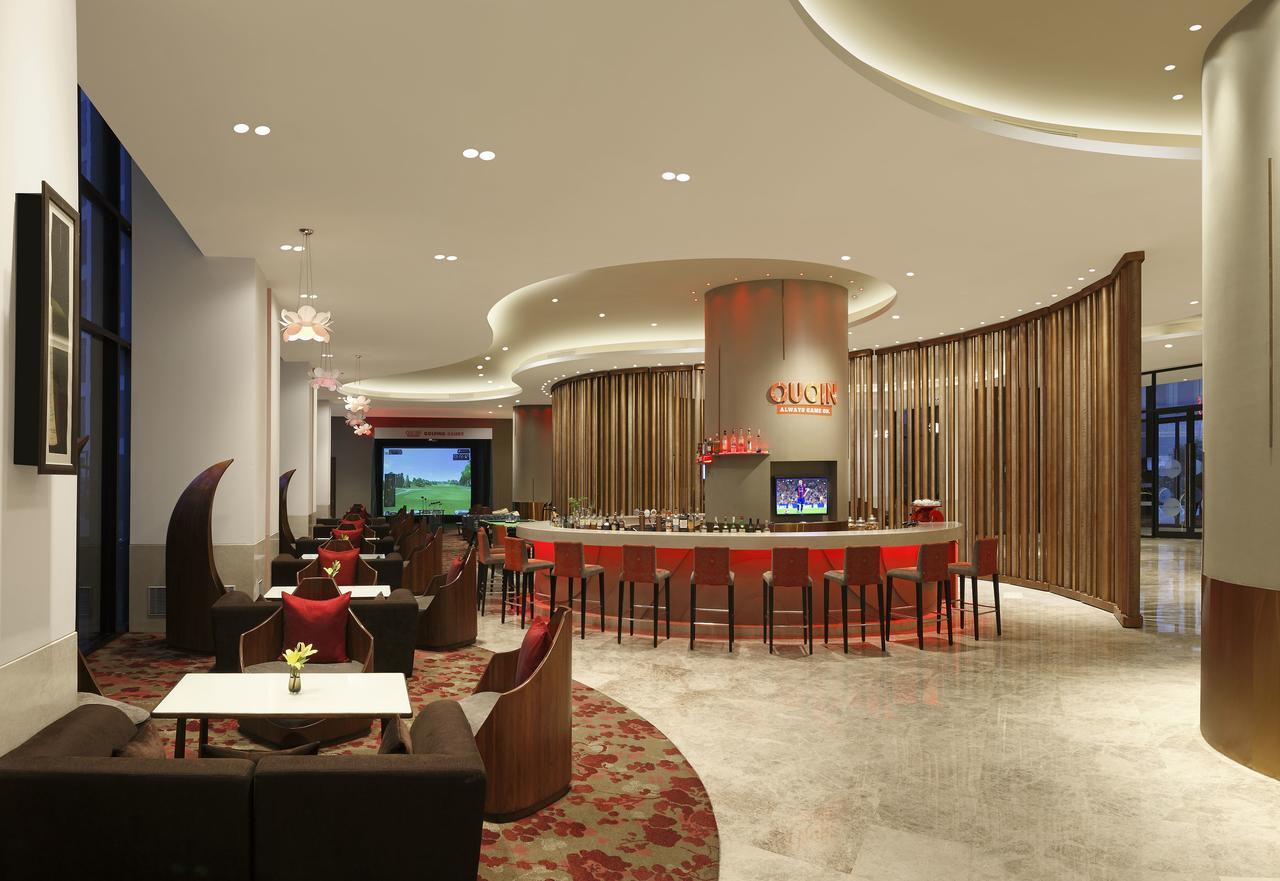 Novotel New Delhi Aerocity Buitenkant foto The photo shows a modern and stylish restaurant or lounge area. There is a curved bar area at the center, featuring a large countertop and seating arrangements. The decor includes elegant lighting fixtures and soft seating in warm tones. The space ha