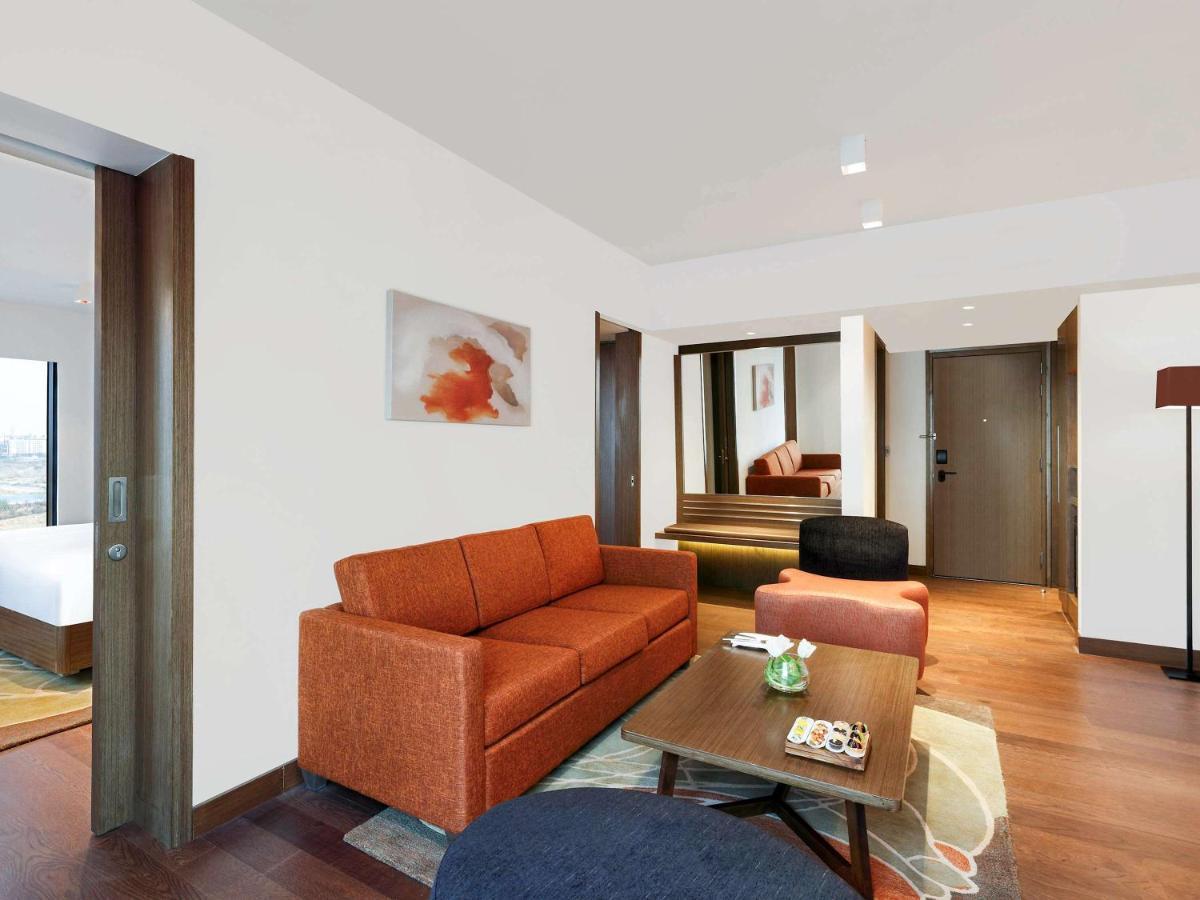 Novotel New Delhi Aerocity Buitenkant foto The photo shows a modern hotel room or suite featuring a spacious living area. There is an orange couch in the center, accompanied by a round blue chair and a coffee table with decorative items on it. The decor includes a piece of wall art, and the r